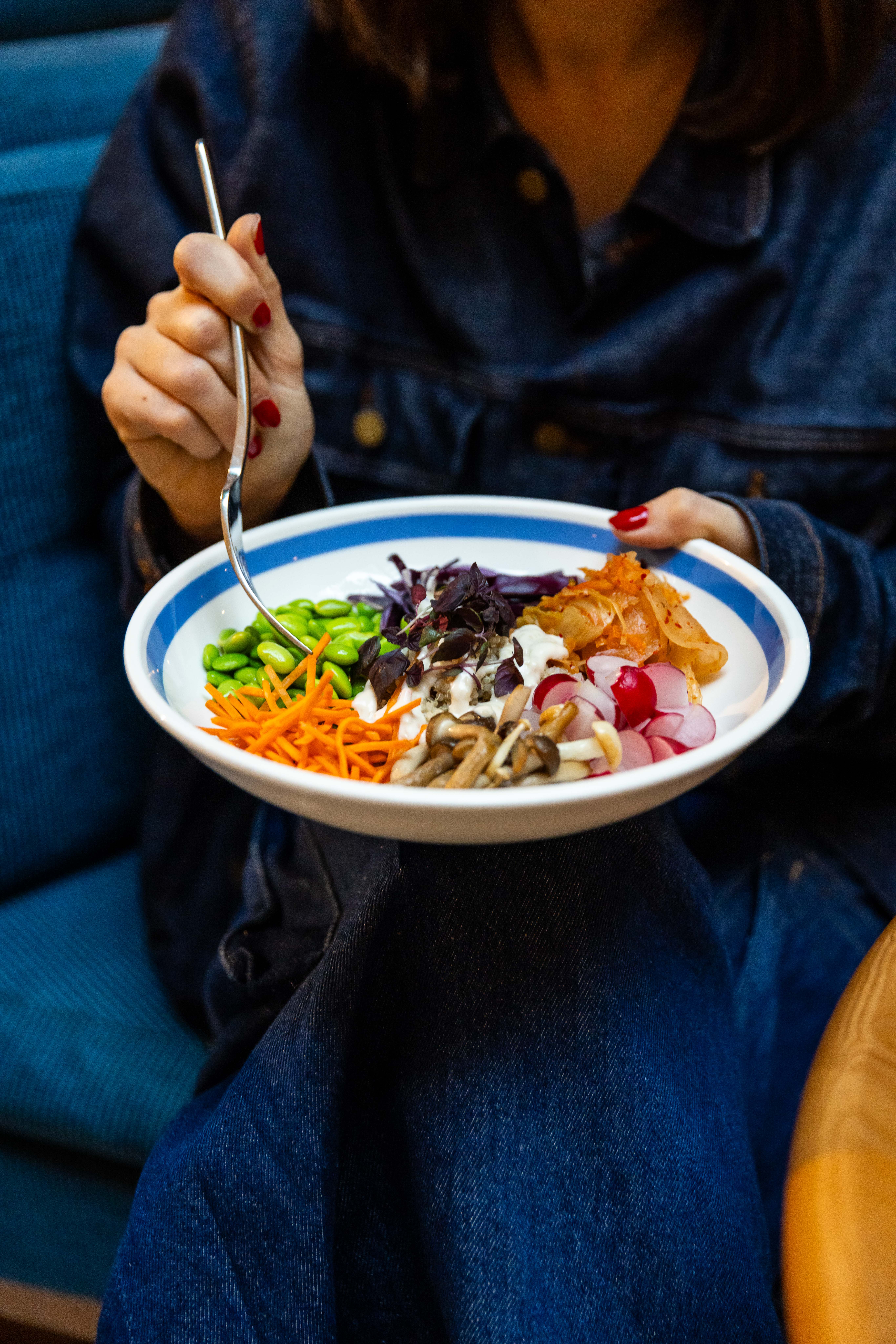 A bowl filled with fresh vegetables, edamame, kimchi, and mushrooms being held by a woman at Celia restaurant in Amsterdam.
