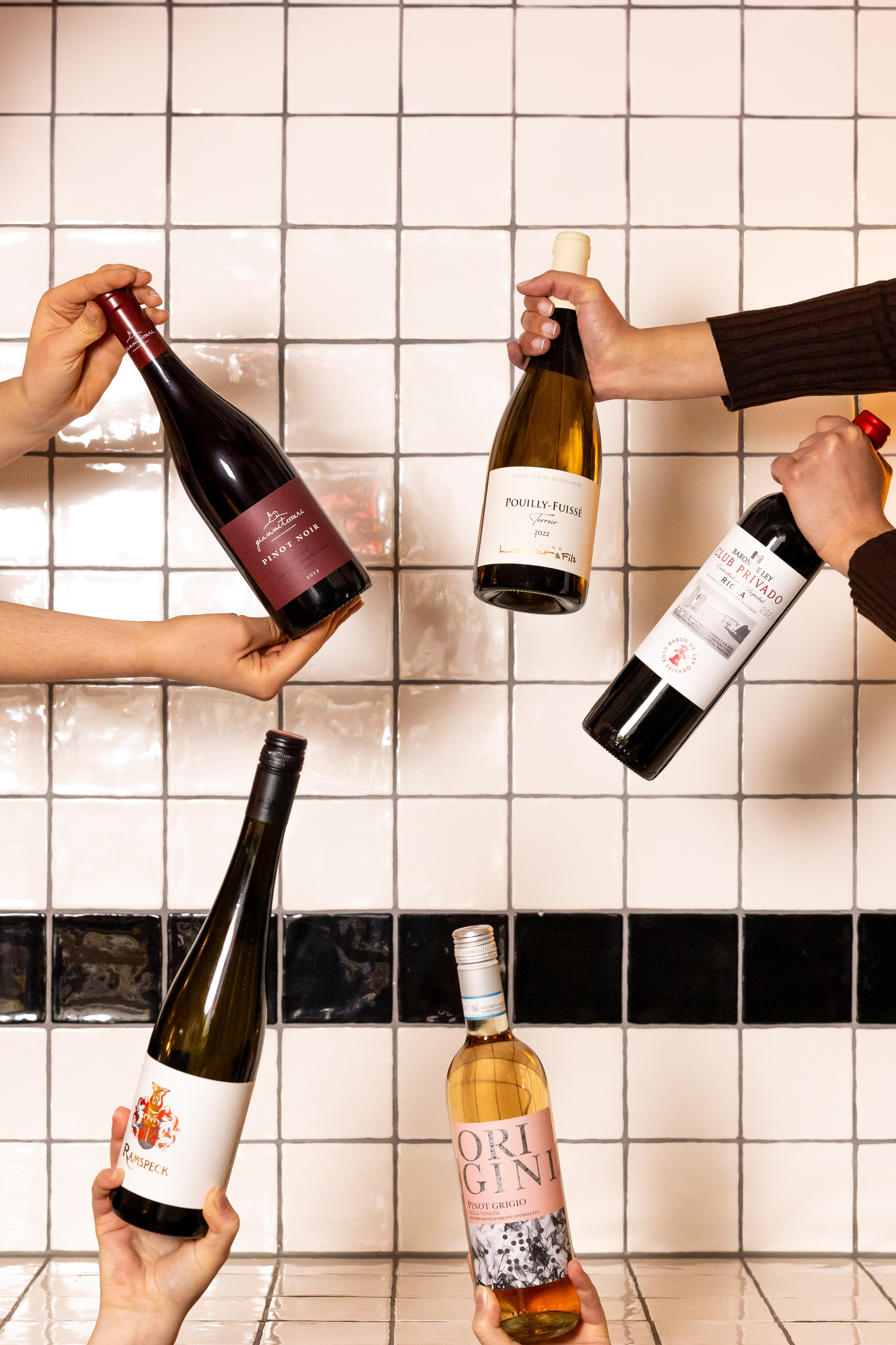 Multiple bottles of wine held up, including red, white, and rosé varieties with a white tiled background at Celia Amsterdam.