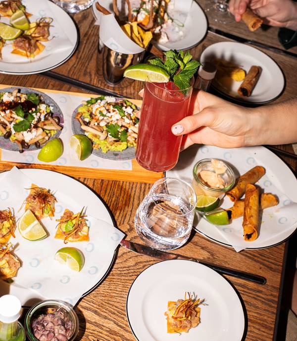 A woman holding a refreshing drink in one hand while surrounded by tacos, sauces, and crispy snacks at Taco Tuesday at Celia.
