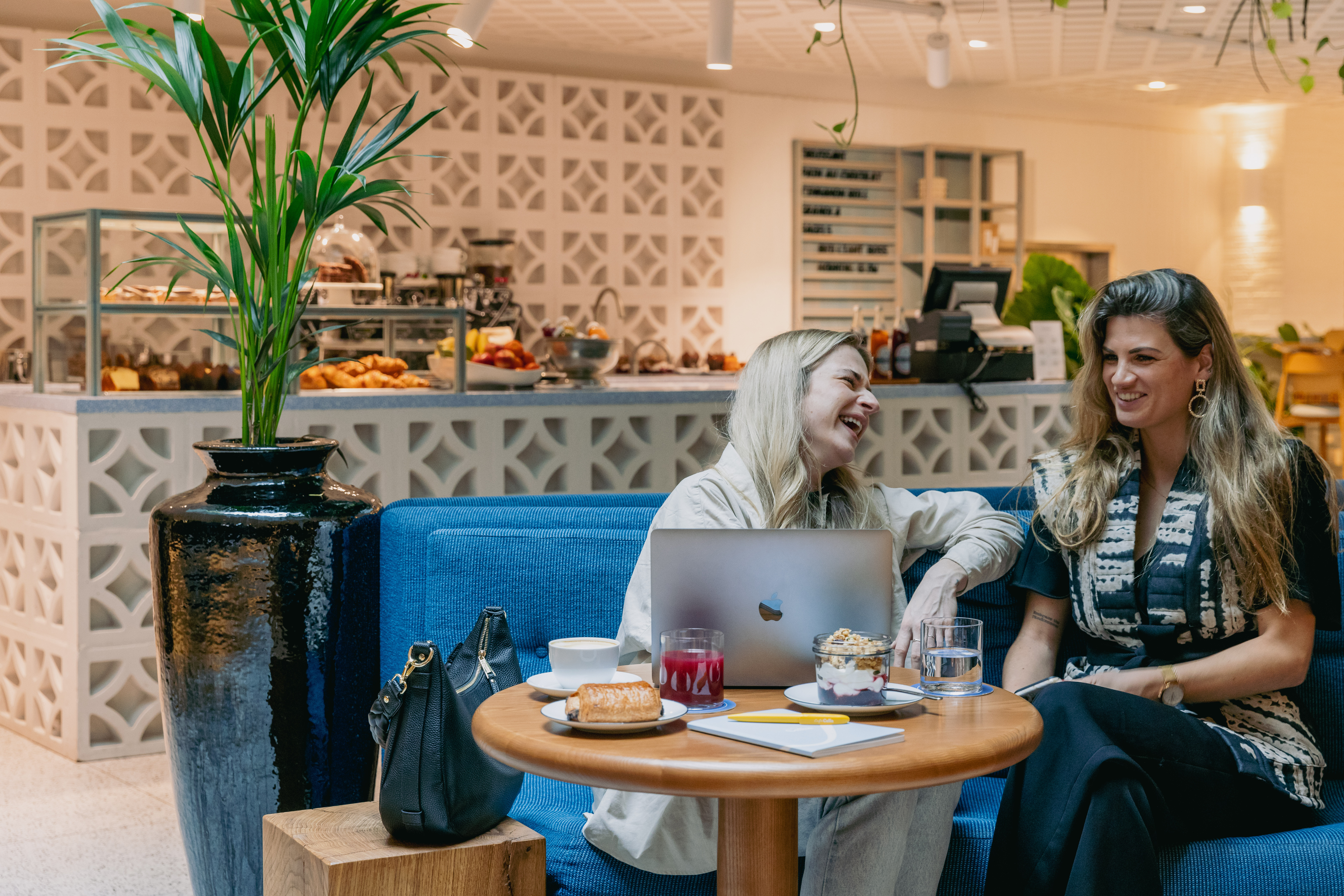 Two women laughing and enjoying drinks while sitting at a table with a laptop and food at Celia.
