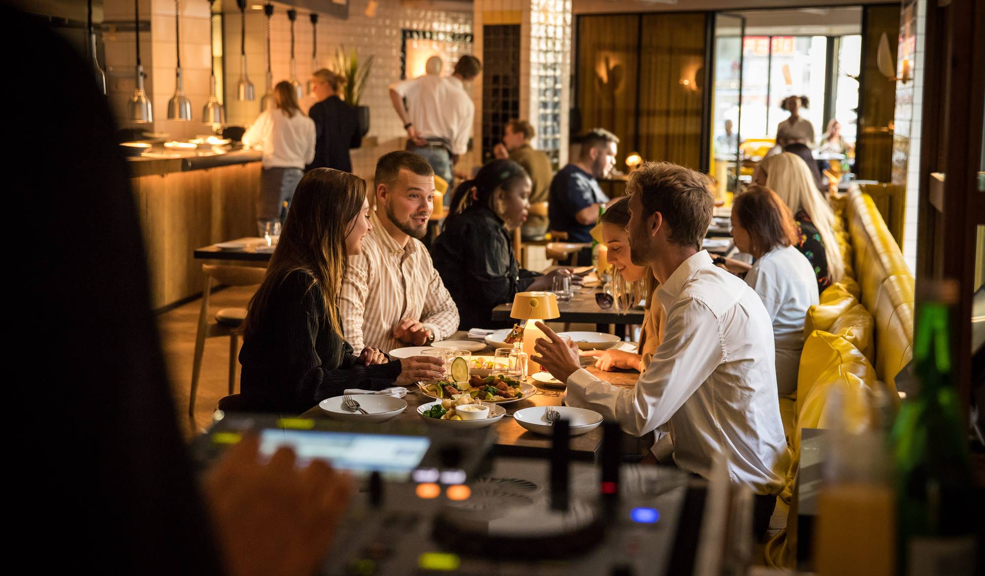 A DJ booth is visible in the foreground while diners chat and enjoy food at Celia's vibrant restaurant in central Amsterdam.